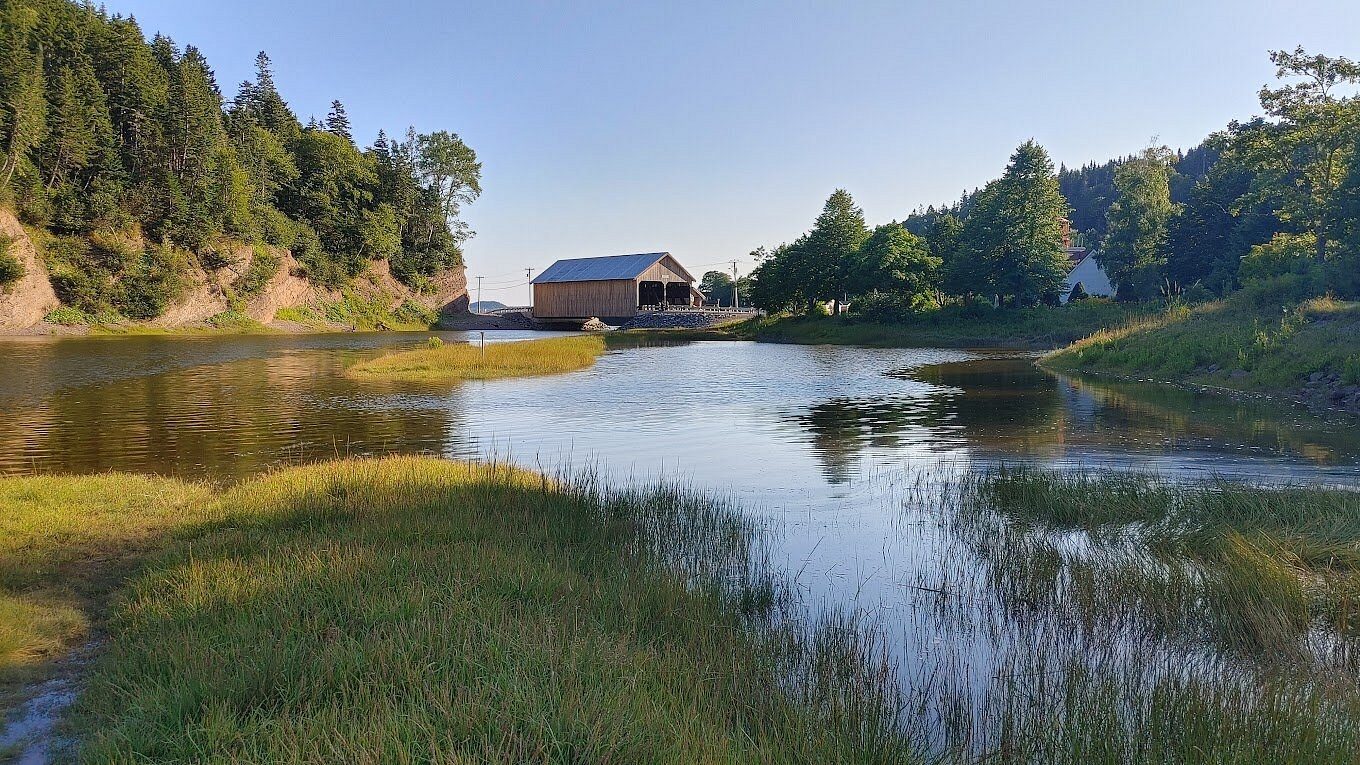 A tidal river and grassy bank beneath forested hills with a boathouse beyond