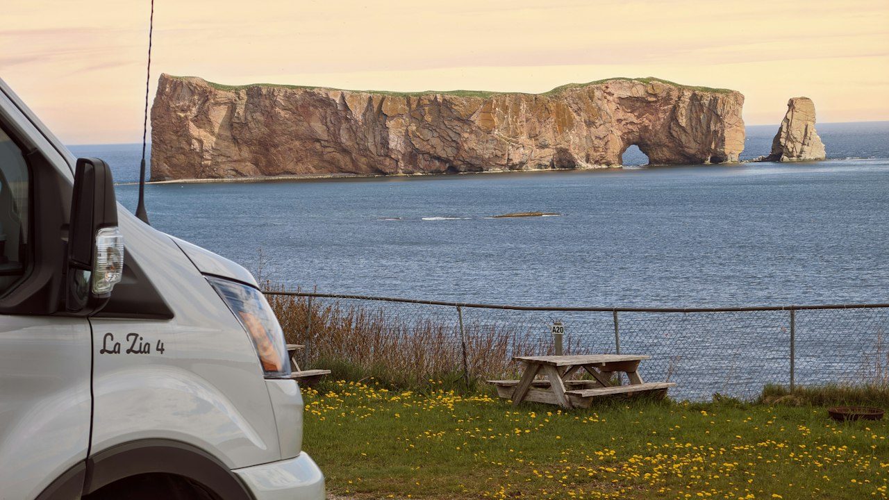 A camper van parked beside a picnic table with Percé Rock rising from the Gulf of St. Lawrence at sunset